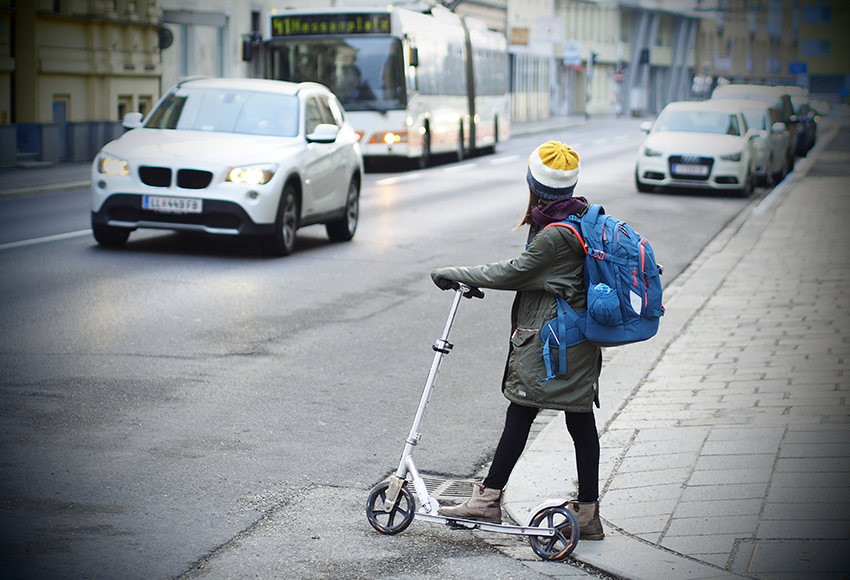 Ein Mädchen steht mit ihrem Roller am Rand einer Straße und beobachtet den Verkehr.