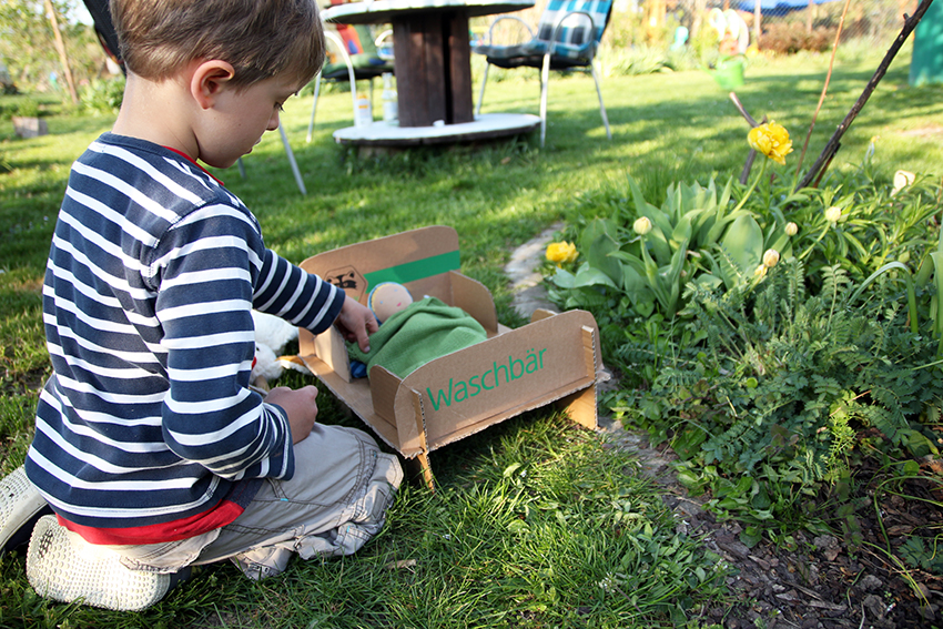 Ein kleiner Junge sitzt im Garten und spielt mit dem Puppenbett aus Karton.