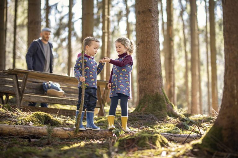 Zwei Kinder entdecken gemeinsam den Wald und seine Waldtiere, ihr Vater ist im Hintergrund zu sehen.