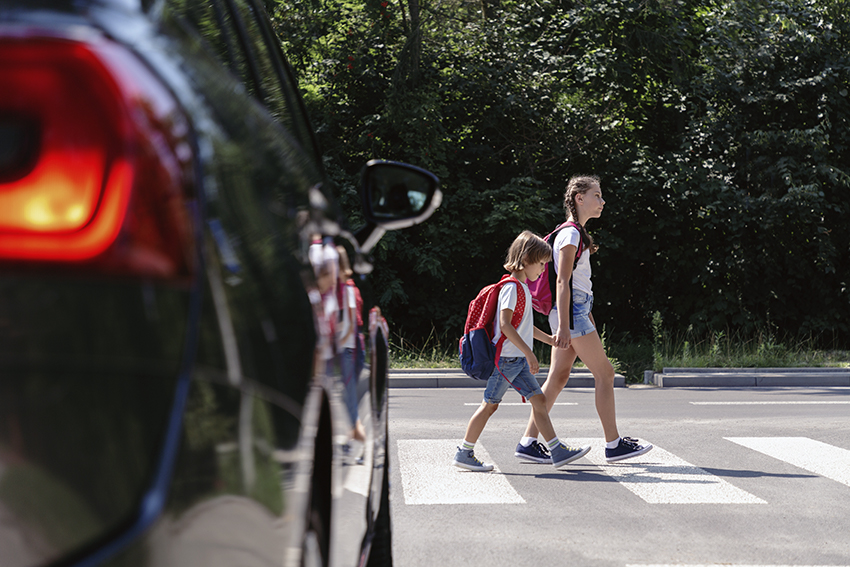 Zwei Schulkinder überqueren einen Zebrastreifen, ein Auto hat vor dem Zebrastreifen angehalten.