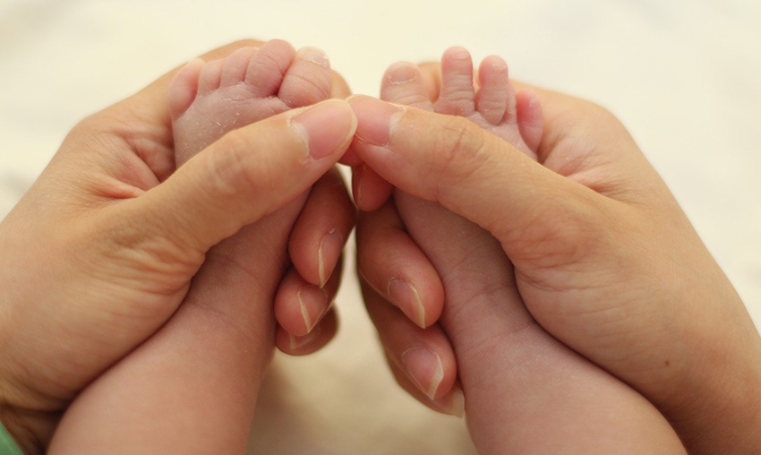 Die beiden Hände halten die Babyfüßchen nebeneinander, deutlich sichtbar ist die trockenere Babyhaut.