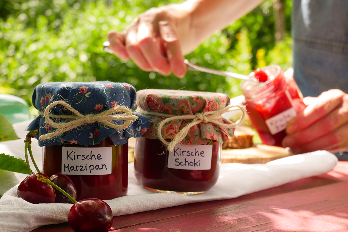 Mehrere Gläser mit Kirschmarmelade stehen auf einem Gartentisch, im Hintergrund wird ein Brot mit Marmelade beschmiert.