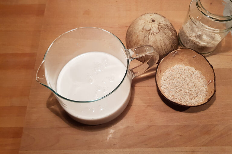 In der Glaskanne sieht man Kokosmilch, in den aufgeschlagenen Nusshälften die Kokosraspel.