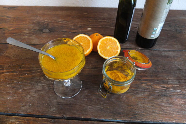 Der Kurkuma-Orangen-Shot steht in einem hübschen Glas mit einem Löffel darin auf einem Holztisch.