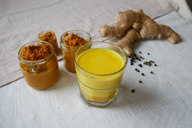 Neben dre kleinen Einmach-Gläaser befüllt mi orangener Paste, steht ein Glas mit gelber Milch und eine Ingwerwurzel.