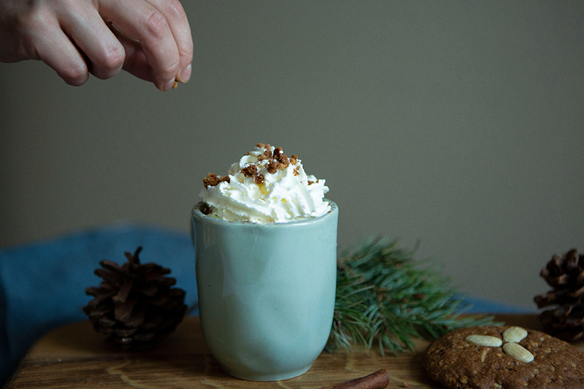 Der Lebkuchen-Latte steht in einer Tasse bereit, auf dem Getränk häuft sich Schlagsahne, die gerade mit Lebkuchen-Bröseln verfeinert wird.