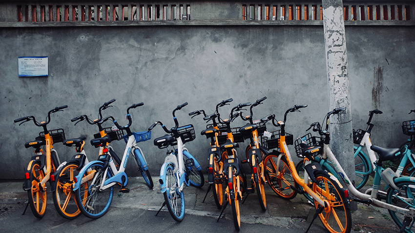 Teil der Sharing Economy sind Leihfahrräder, die hier in orange und blau vor einer grauen Hauswand stehen.