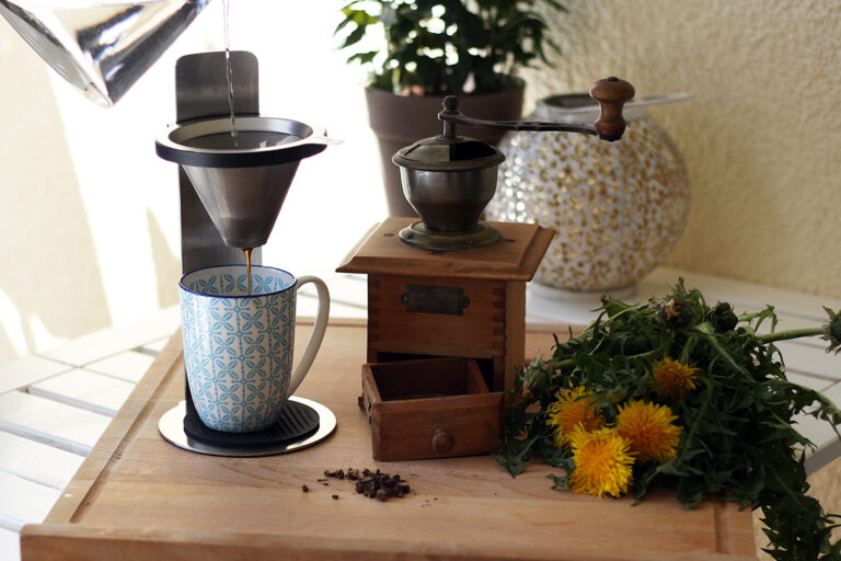 Löwenzahn-Kaffee wird frisch aufgebrüht und in eine Tasse gegossen, daneben steht eine alte Kaffeemühle und Löwenzahn liegt aus.
