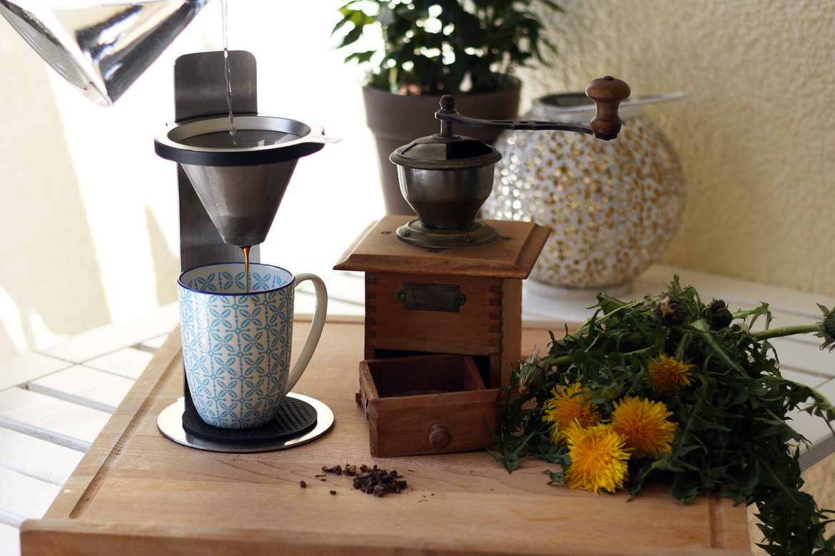Löwenzahn-Kaffee wird frisch aufgebrüht und in eine Tasse gegossen, daneben steht eine alte Kaffeemühle und Löwenzahn liegt aus.
