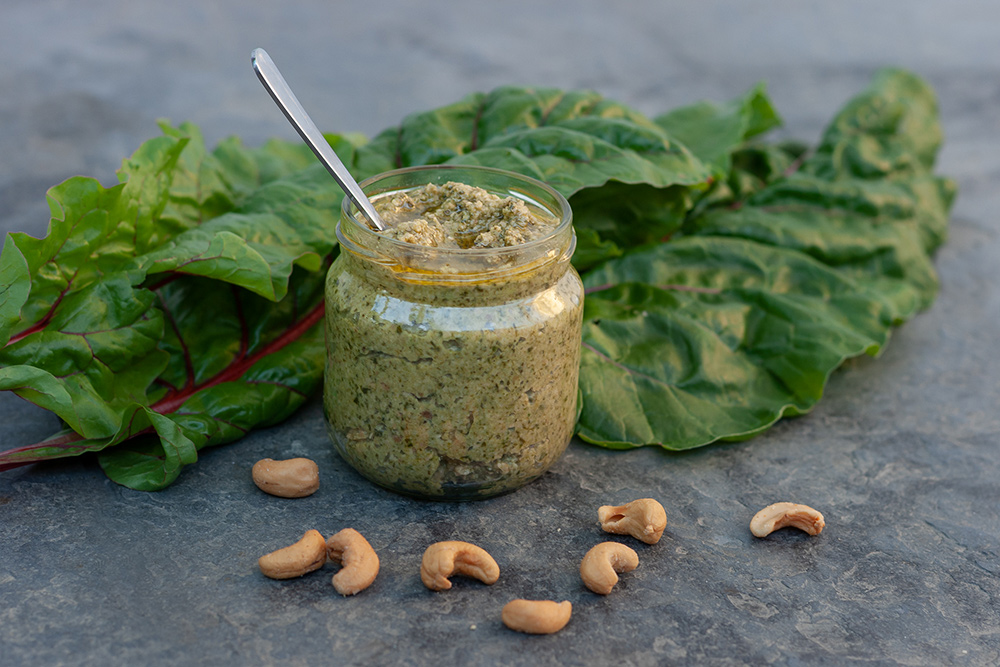 Ein geöffnetes Glas mit grünem Pesto steht vor Mangoldblättern mit rotem Stiel.