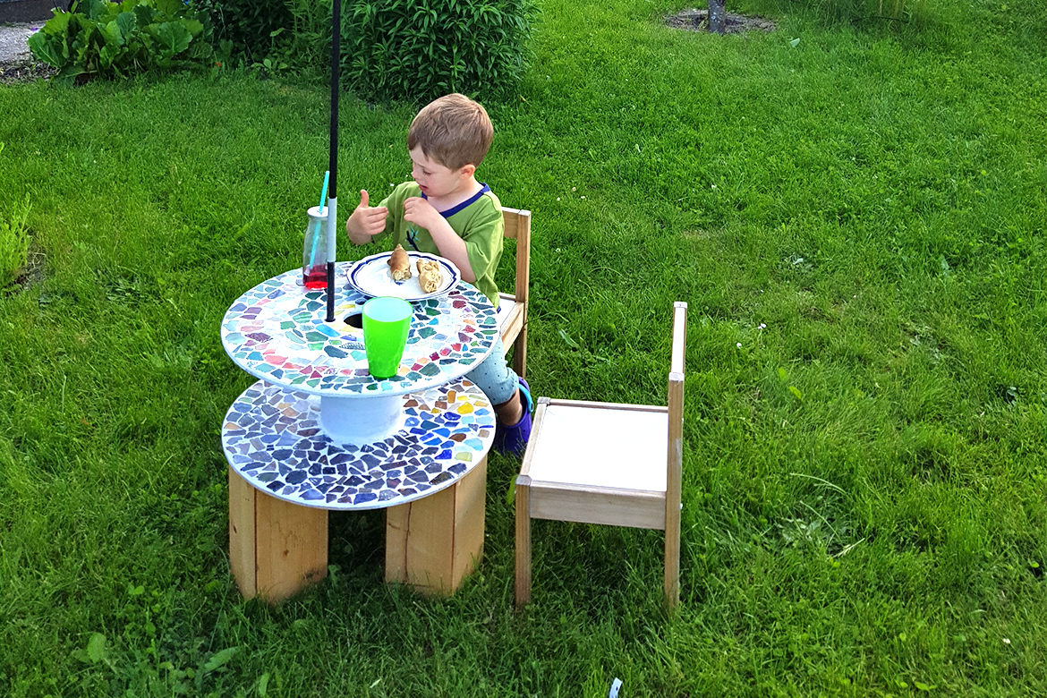 Der kleine Junge sitzt am Tischchen im Garten, neben ihm ist noch ein Stühlchen frei.