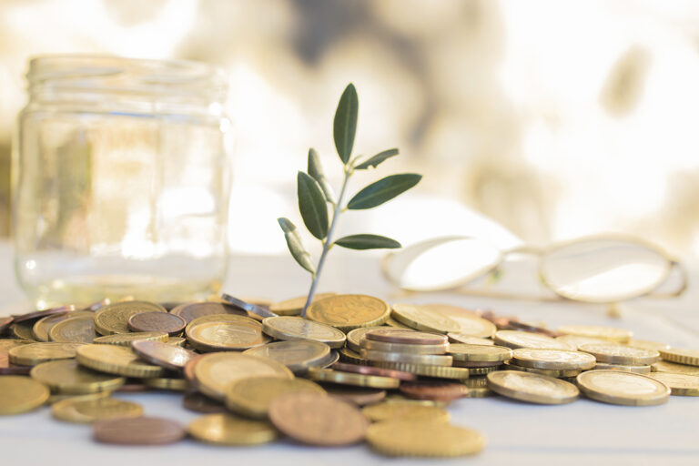Münzen liegen auf dem Tisch und aus der Mitte des Geldhaufens wächst eine kleine Pflanze.