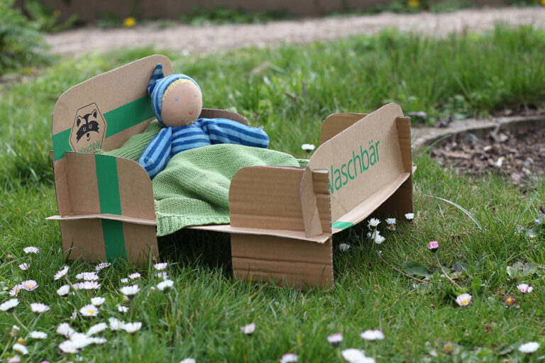Ein Puppenbett aus Karton mit einer Puppe darin steht auf dem Rasen.