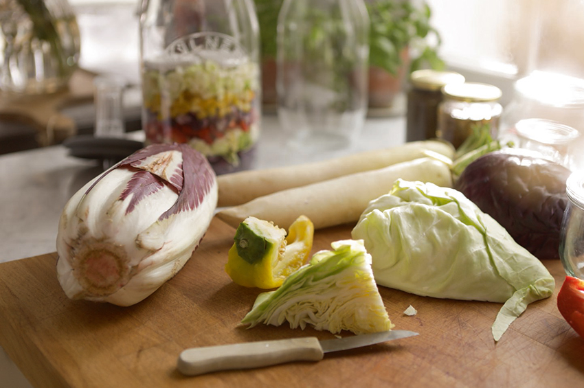 Radiccio und Eisbergsalat liegen auf einem Schneidebrett, im Hintergrund sind weitere frische Zutaten als Schichtsalat in ein Glas gefüllt.