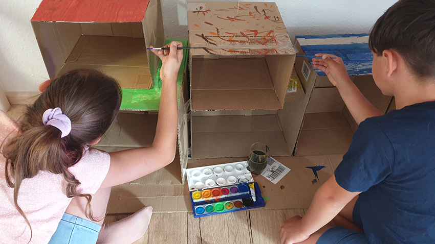 Zwei Kindern basteln mit Karton ein Regal, das sie mit Wassermalfarben verschönern.