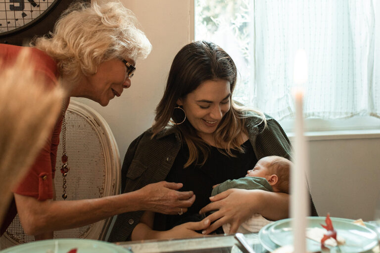 Eine Mutter sitzt mit ihrem Baby im Arm am Tisch in einem Mehrgenerationenhaus, eine ältere Frau begrüßt das kleine Kind.