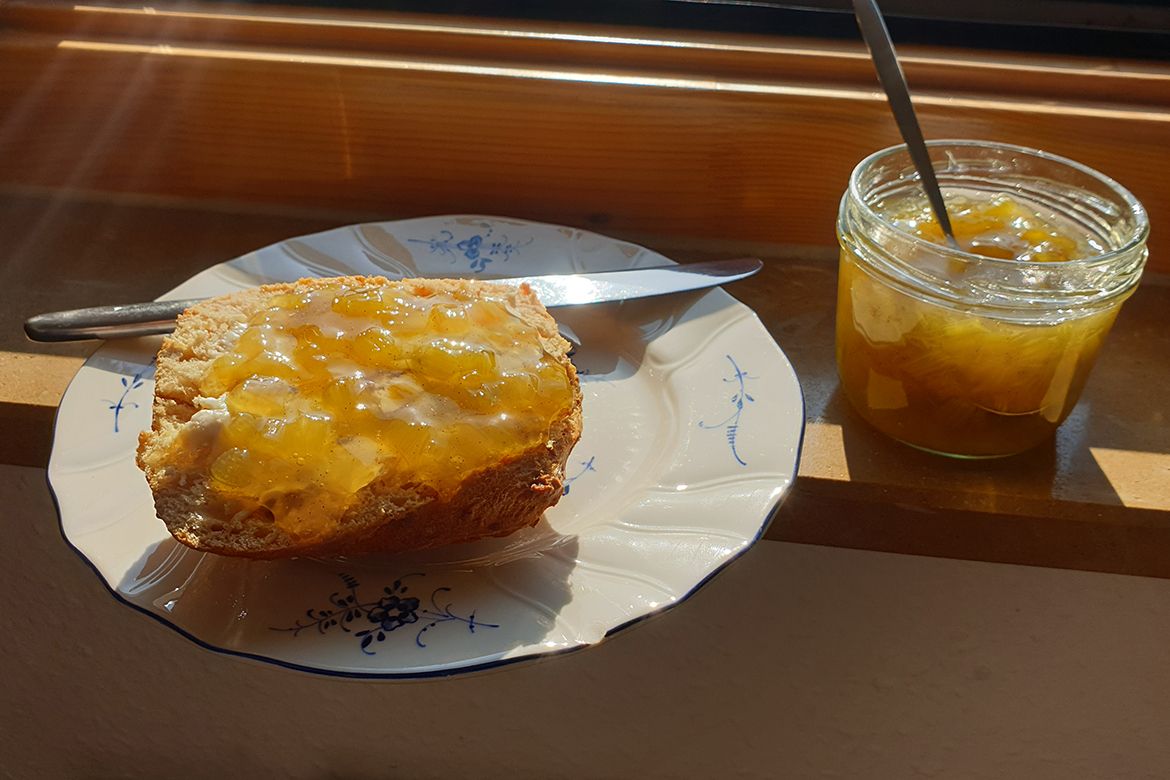 Ein Brötchen mit Rhabarber-Marmelade steht neben dem Marmeladenglas auf einer sonnigen Fensterbank.