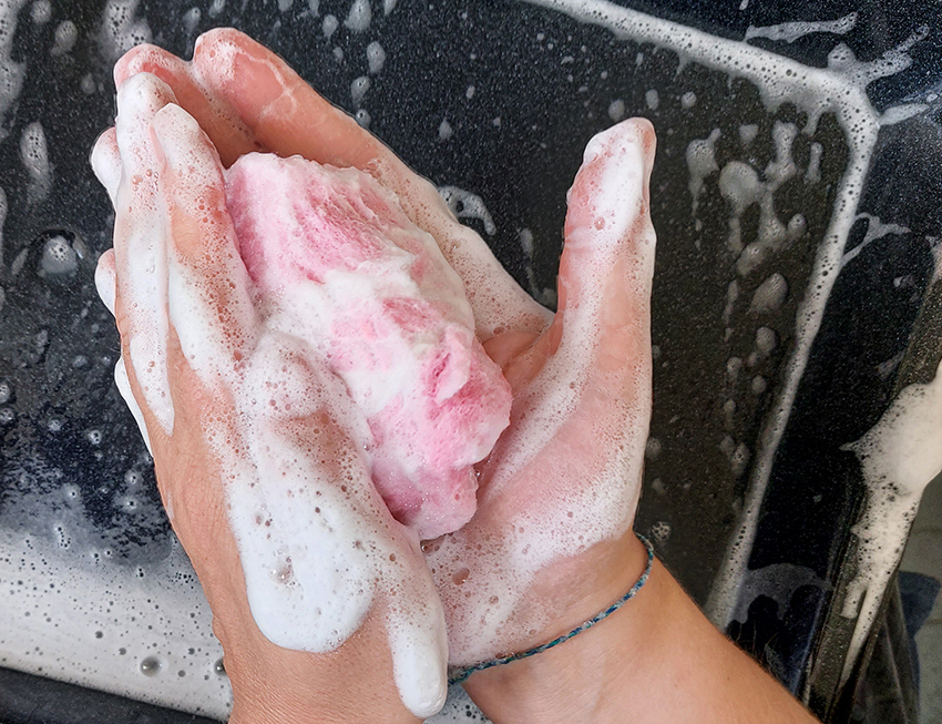 Zwei Hände halten eine rose gefilzte Seife in den Händen über einen Backblech, das Seife filzen schäumt kräftig.