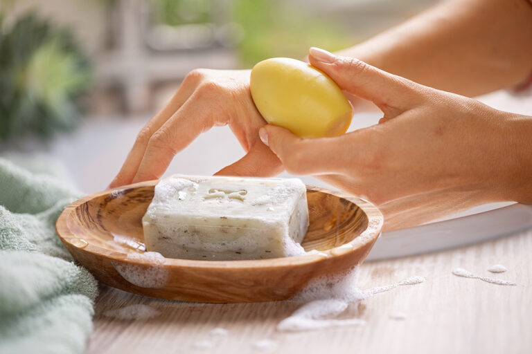 Eine Seife liegt in einer Schale aus Holz, dahinter werden Hände mit einer eiförmigen Handcreme gepflegt.