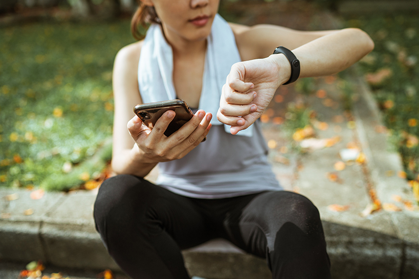 Frau sitzt auf dem Bürgersteig und kontrolliert mittels digitaler Selbstvermessung ihre Fitnessdaten auf Smartphone und Fitnessuhr.