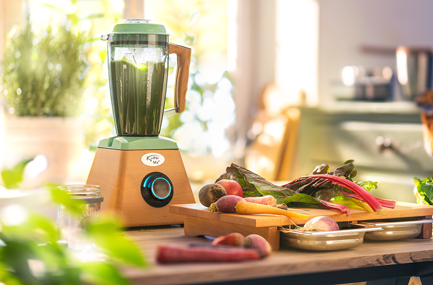 In einer Küche steht ein Mixer, der einen grünen Smoothie zubereitet – auf dem Schneidebrett daneben liegt restliches Obst.