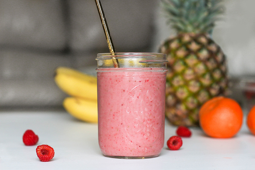 Ein Glas mit einem rosafarbenen Smoothie, Im Hintergrund sind Bananen, Orangen und eine Ananas zu sehen.