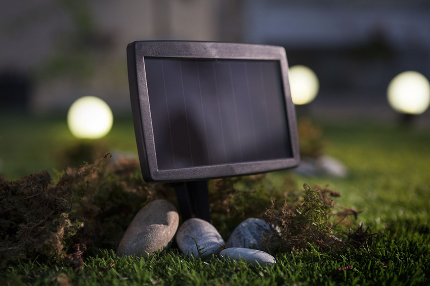 Ein Solarpanel steckt im Rasen, im Hintergrund leuchten die Gartenlampen in der Abenddämmerung.