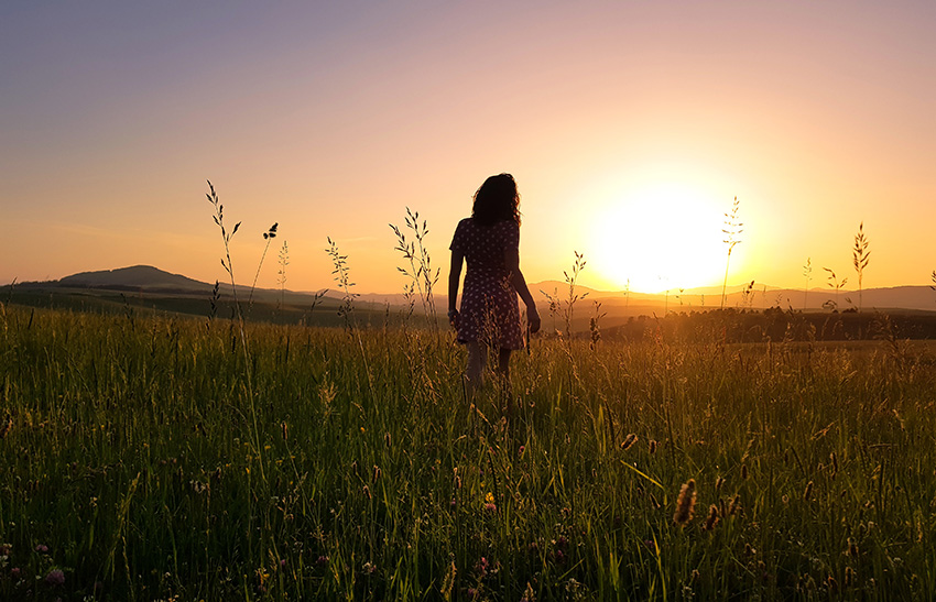 Eine Frau im Kleid steht auf einer Wiese und betrachtet den Sonnenuntergang.
