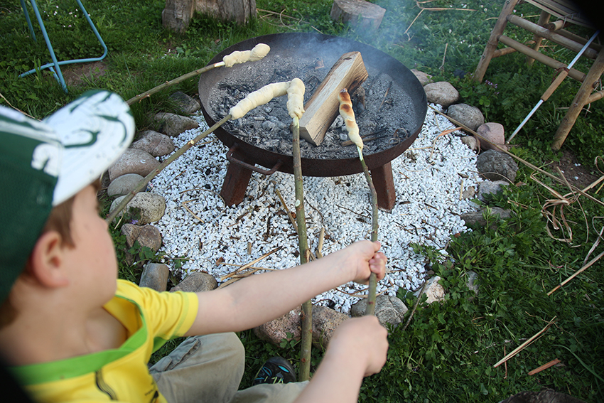 Ein kleiner Junge sitzt vor der Feuerschale und hält sein Stockbrot über das Feuer.