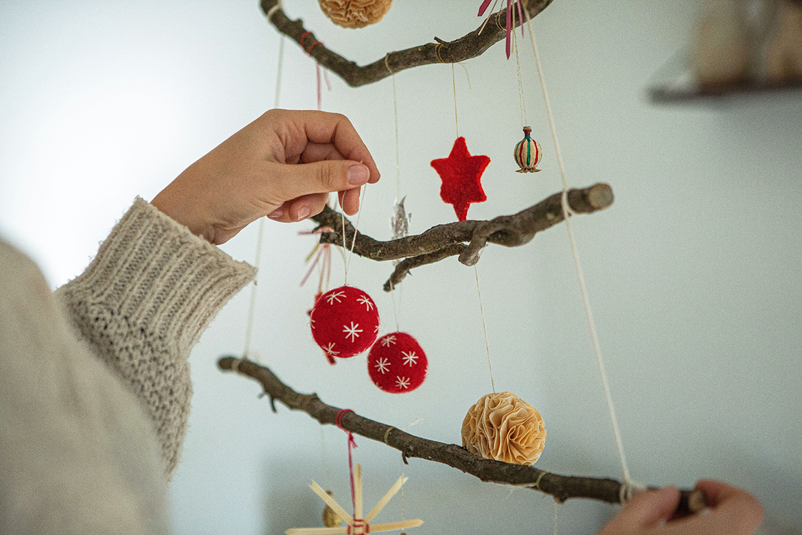 Am Weihnachtsbaum aus Ästen hängt bereits Baumschmuck, eine Hand hängt gerade eine rote Filzkugel an einen Ast.
