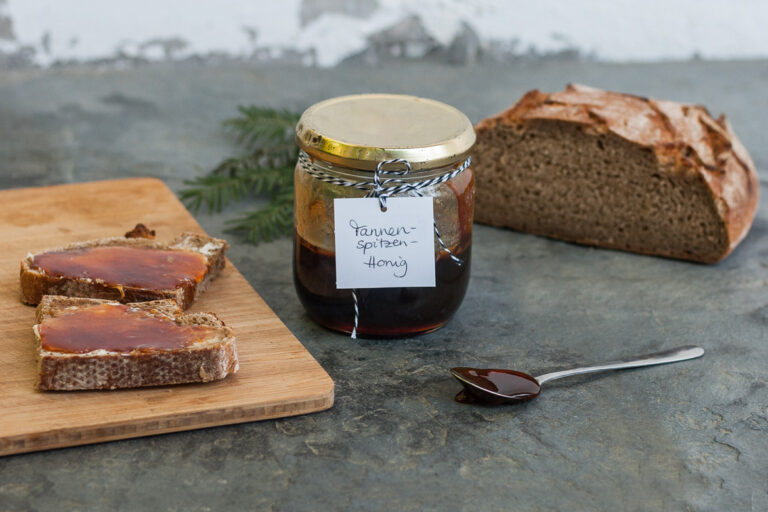 Ein Glas Tannenspitzenhonig, daneben Brotscheiben, ein Laib Brot, ein Löffel und ein Tannenzweig