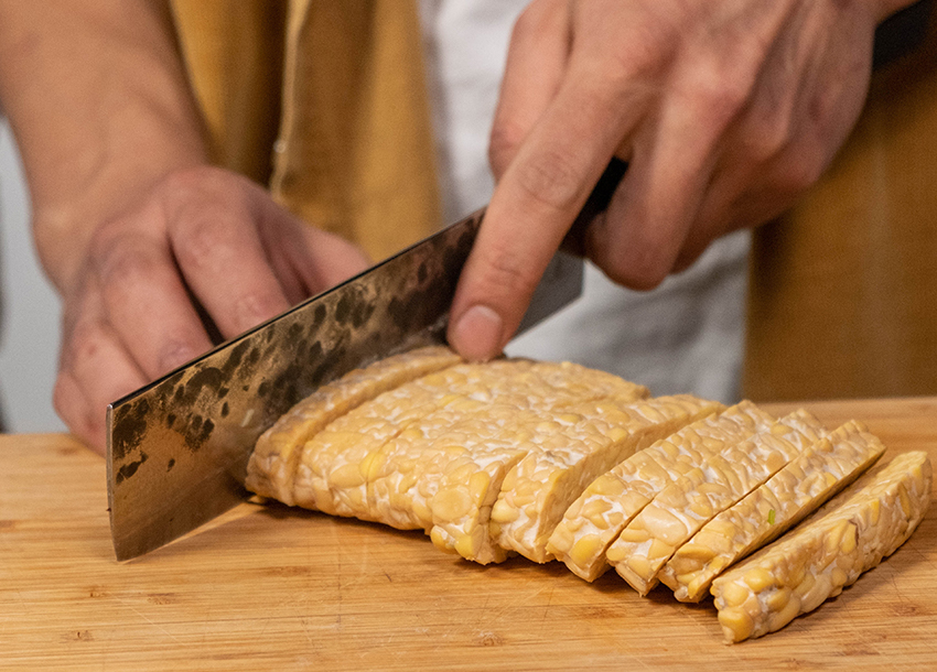 Tempeh liegt auf einem Holzbrett und wird mit einem großen Messer in Scheiben geschnitten.