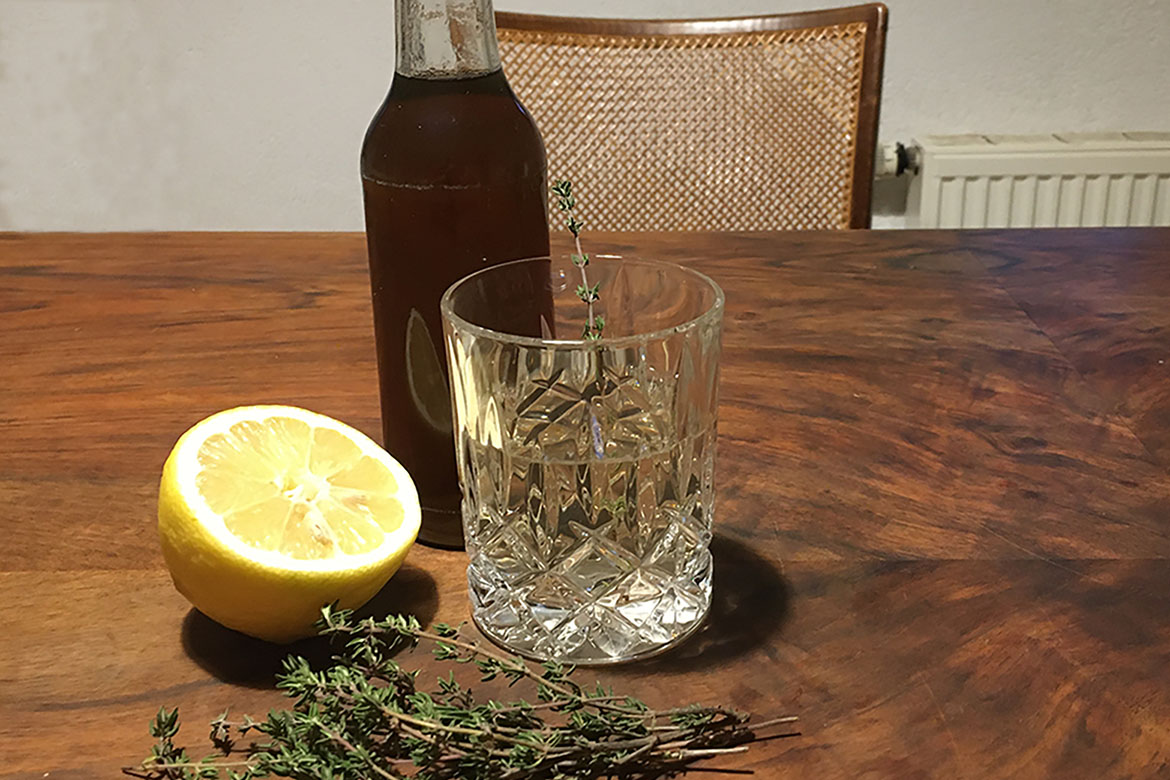 In Flasche abgefüllter Sirup neben einem Wasserglas, einer angeschnittenen Zitrone und einem Ästchen Thymian.