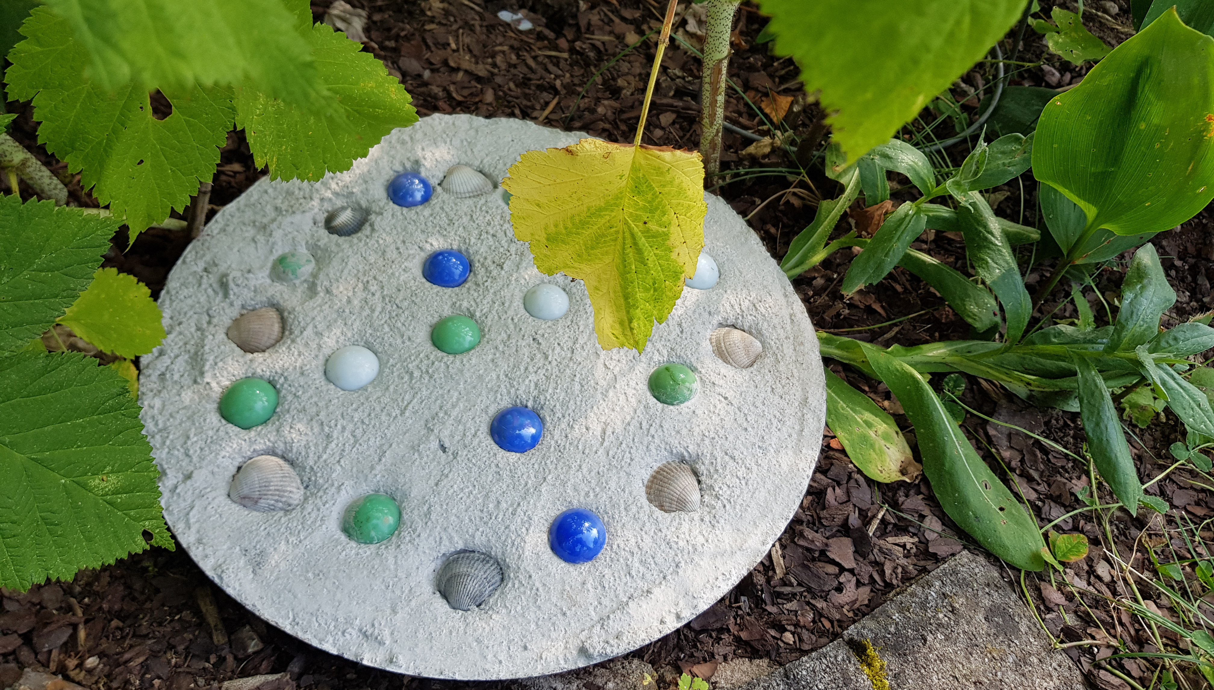 Ein runder Trittstein mit blauen, grünen, weißen Murmeln und Muscheln.