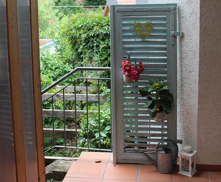 Ein großer Fensterladen wurde mit blauer Kreidefarbe angestrichen und steht dekorativ neben dem Hauseingang.