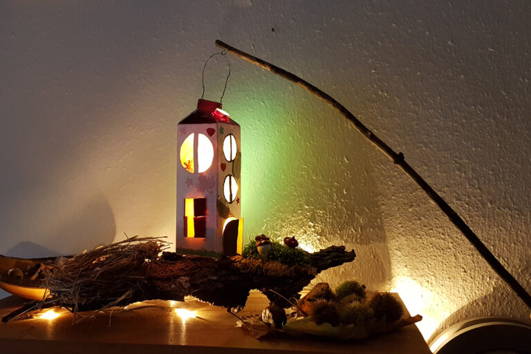 Die hohe Upcycling-Laterne leuchtet vor der weißen Wand.