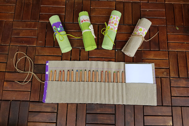 Ein ausgerolltes Upcycling-Mäppchen Mäppchen mit Buntstiften und Papier befüllt liegt vor vier aufgerollten bunten Mäppchen.