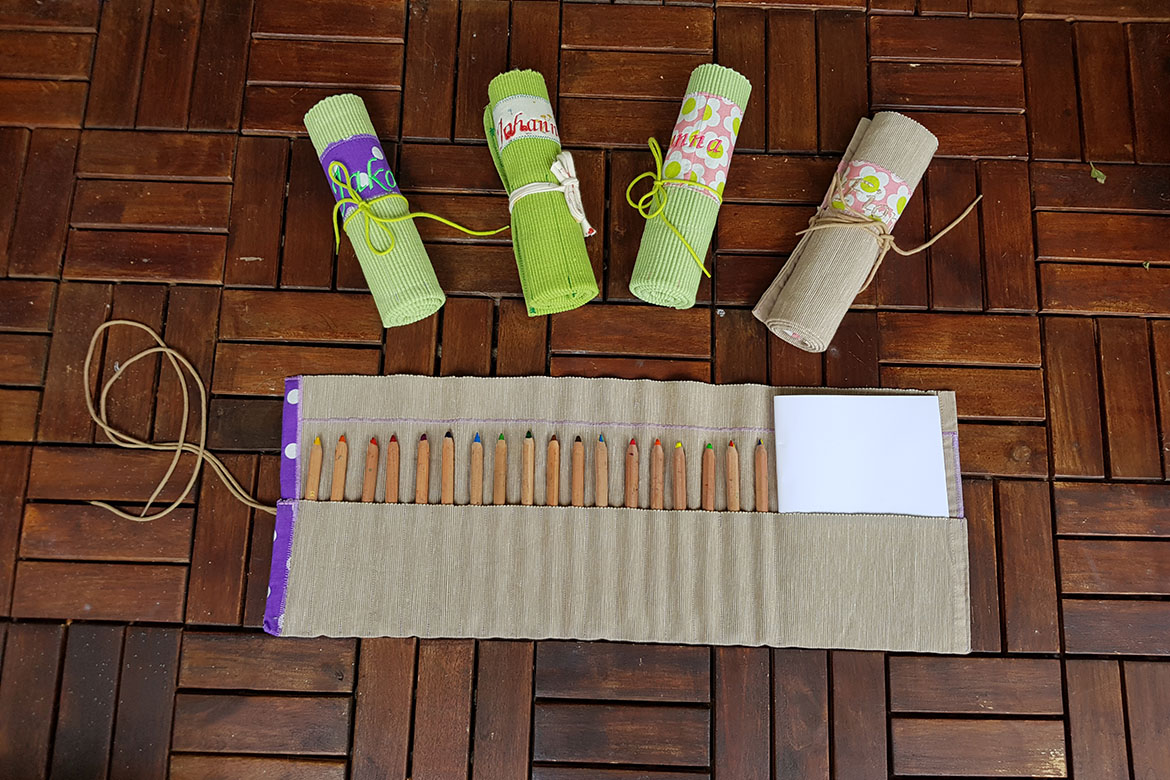 Ein ausgerolltes Upcycling-Mäppchen Mäppchen mit Buntstiften und Papier befüllt liegt vor vier aufgerollten bunten Mäppchen.