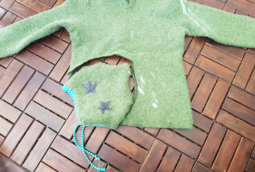 Der grüne Pullover liegt ausgebreitet da, die Upcycling-Walkmütze liegt dort, wo der Stoff ausgeschnitten wurde.