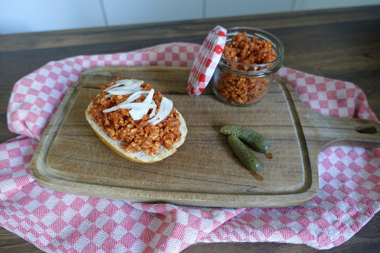 Auf dem Holzbrett liegt ein Brötchen mit Mett und Zwiebeln belegt, nebenan zwei kleine Essiggurken und ein Glas mit veganem Mett.