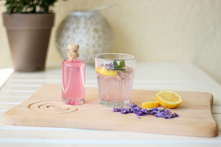 In einer Flasche abgefüllter Veilchen-Sirup steht neben einem Glas mit Sirup auf einem Schneidebrett aus Holz.