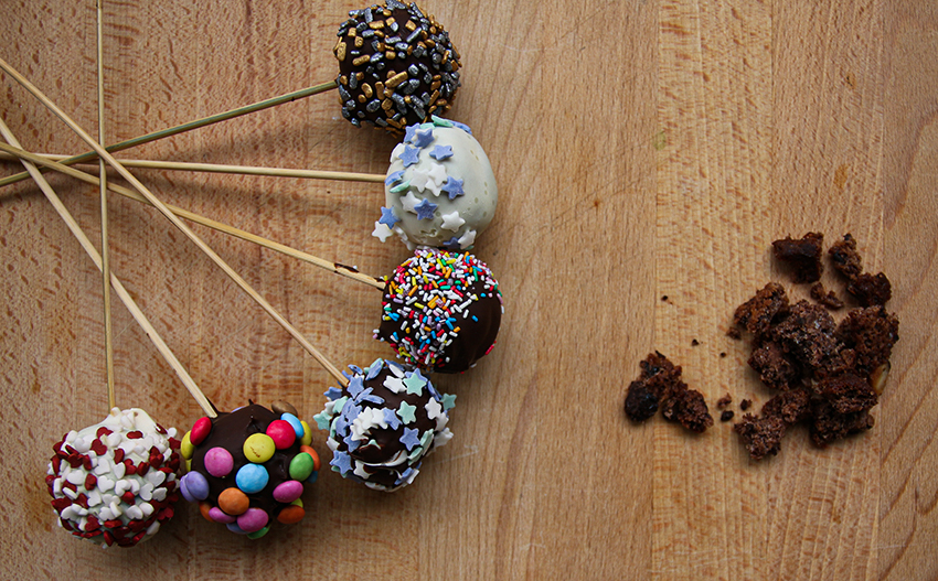 Verzierte Cake Pops liegen aufgefächert auf einem Holzbrett neben Kuchenbröseln.