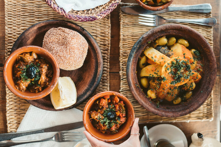 Auf einem Tisch steht eine Tajine mit Gemüse sowie Teller und Schüsseln mit wärmenden Lebensmitteln..