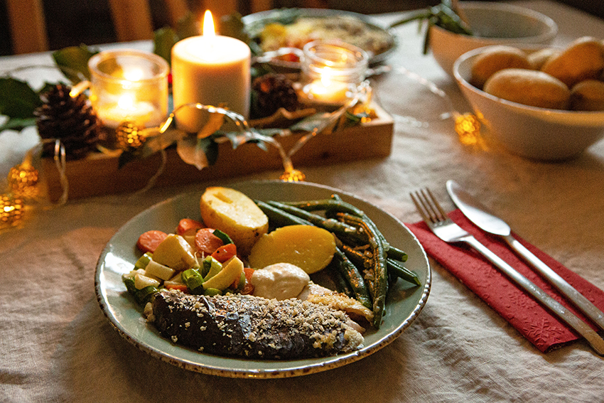 Een stuk kerstkarper wordt geserveerd met aardappelen, bonen en gestoofde groenten op een bord dat op een tafel met kaarsen staat.