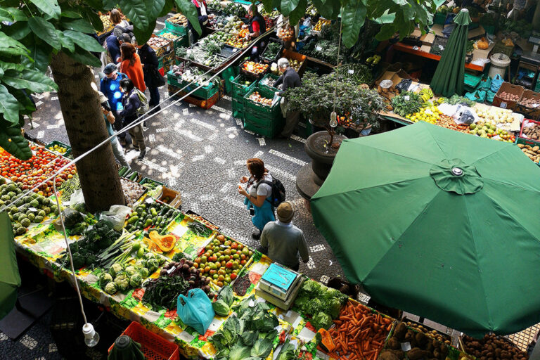 Auf einem Markt wird an bunten Ständen Obst und Gemüse nach dem Saisonkalender angeboten, Menschen gehen auf diesem Markt einkaufen.