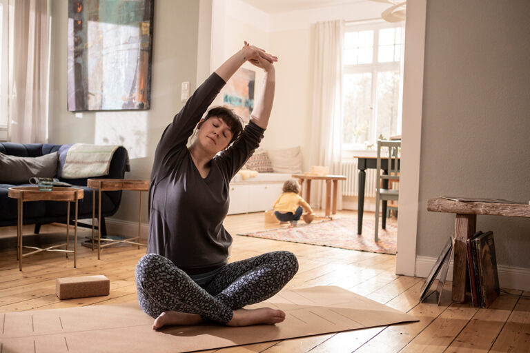 Frau sitzt auf einer Yogamatte aus Kork im Wohnzimmer und dehnt ihre Arme seitlich über den Kopf.