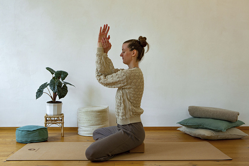 Lea Uhle sitzt auf einem Yogablock aus Kork und führt das Asana Adlerarme durch.