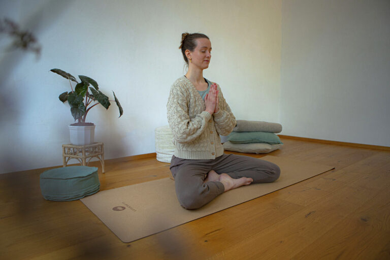 Eine Frau sitzt im Schneidersitz auf einer Yogamatte aus Kork und macht Yoga im Homeoffice, ihre Hände hat sie vor der Brust gefaltet.