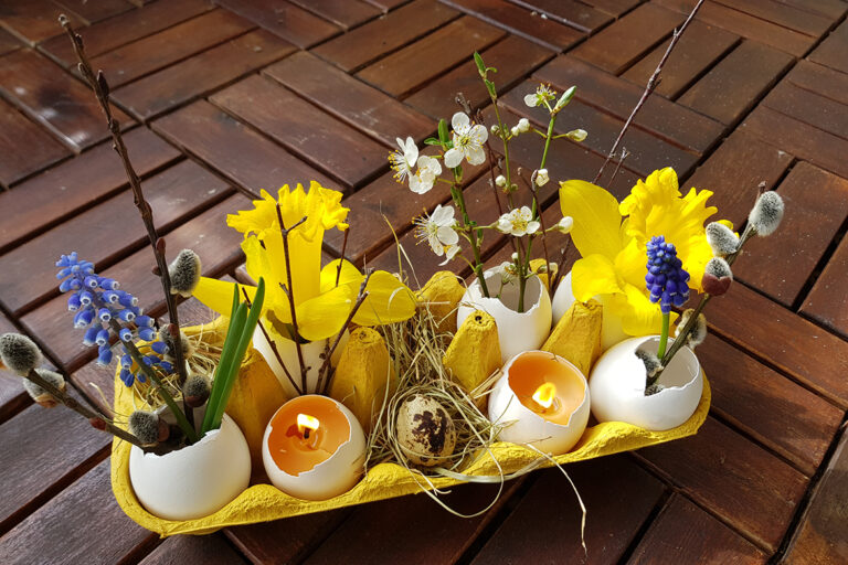 Im gelben Eierkarton sind Ostereier-Kerzen und Blumen drapiert.