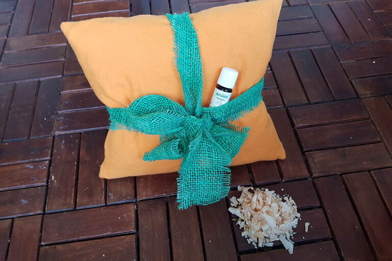 Ein orangenes Kissen ist mit einer grünen Schleife verziert, in der ein Fläschchen Zirbenöl steckt.